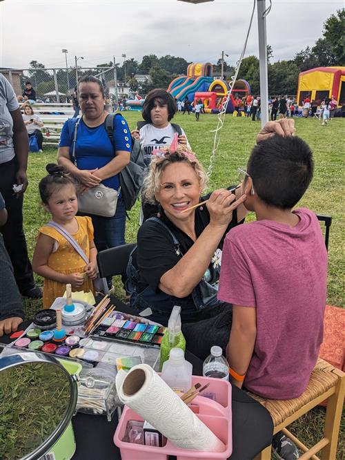 A face painter smiles while painting the face of a child. Other children stand in line.